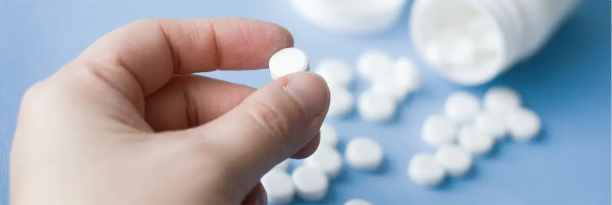 Hand holding a white prescription pill with spilled tablets and an open pill bottle in the background.