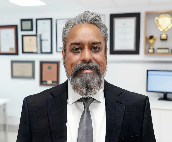 A professional portrait of Dr. Ash Bhatt, M.D., M.R.O., F.A.S.A.M., wearing a black suit and grey tie, standing in an office with medical credentials and awards displayed in the background.