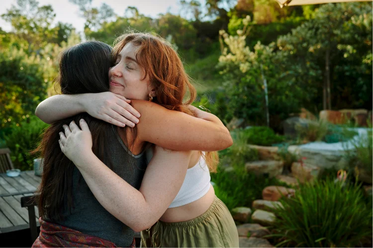 Two women sharing a supportive hug in a lush, private garden setting at a luxury rehabilitation center in Los Angeles.